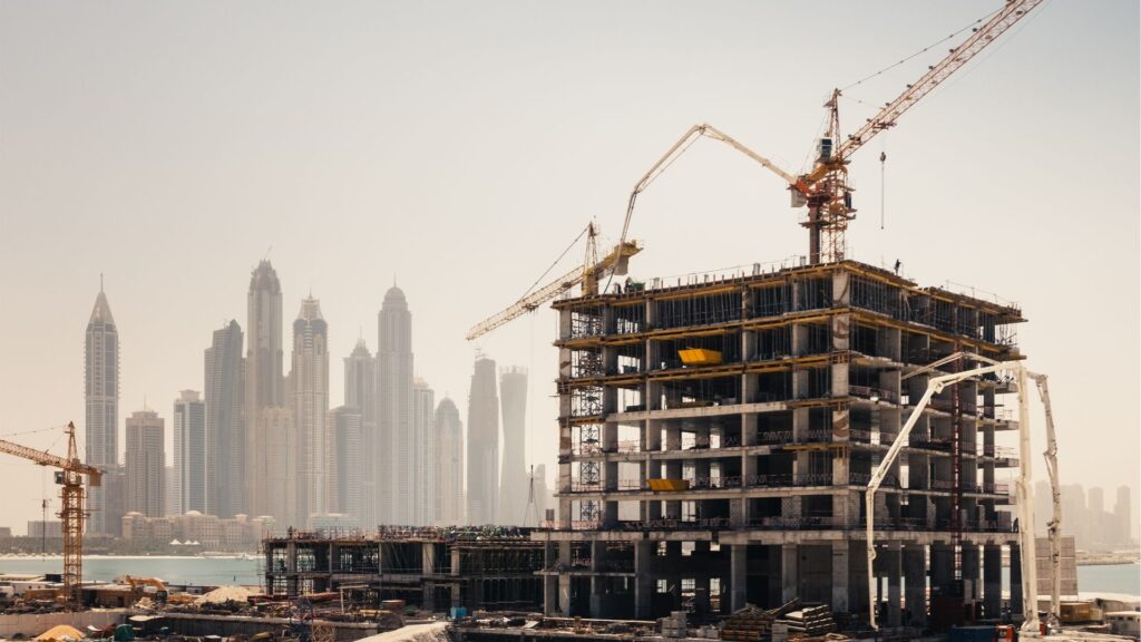 A multi-story building under construction with cranes and scaffolding, set against an urban skyline, highlights why new real estate is so attractive to investors amid tall skyscrapers and a hazy sky in the background.