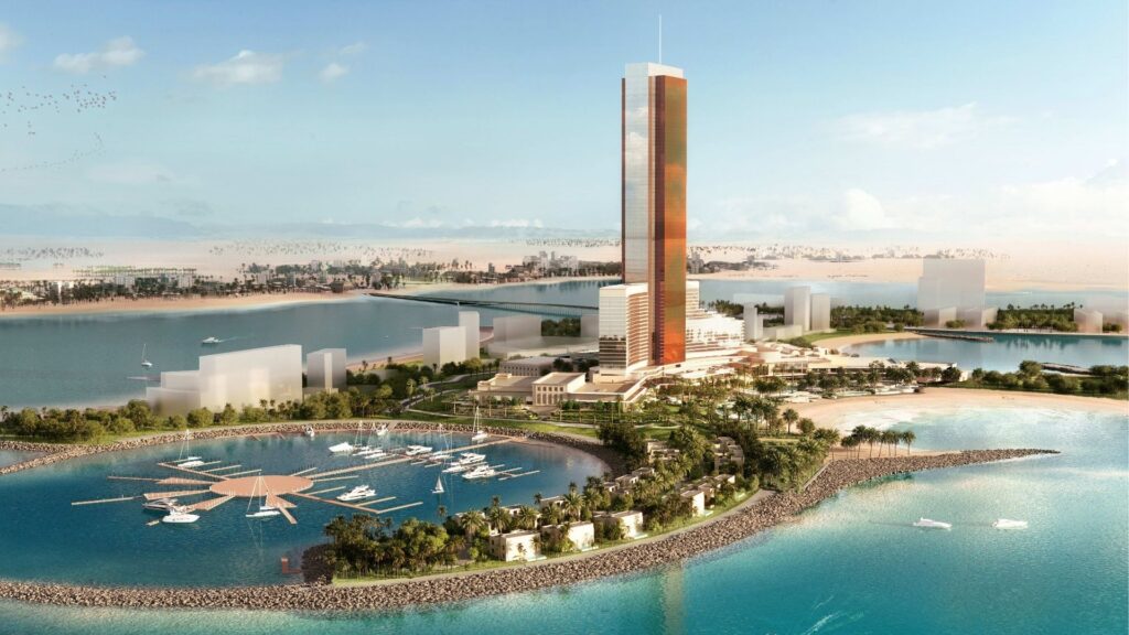 Ein großer moderner Wolkenkratzer ragt auf einer Insel am Wasser in Ras Al Khaimah empor, umgeben von Grünflächen, Kais und Booten – eine Kulisse, die die Frage aufwirft: Sollte man in Immobilien in Ras Al Khaimah investieren? Strände und Ausblicke auf die Wüste runden das Bild ab.