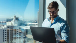 Ein Mann mit Brille und hellblauem Hemd steht an einem großen Fenster in einem Wolkenkratzer und lächelt, während er an seinem Laptop arbeitet – vielleicht recherchiert er gerade zum Thema „Steuern in Dubai: Warum europäische Investoren aktiv werden“. Hinter ihm ist die Stadtlandschaft zu sehen.