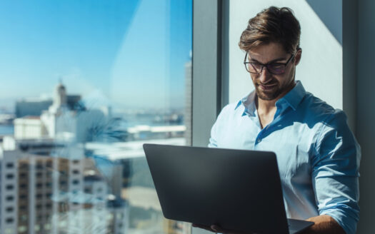 Un homme portant des lunettes et une chemise bleu clair se tient près d'une grande fenêtre dans un gratte-ciel, souriant tout en travaillant sur son ordinateur portable - peut-être en faisant des recherches sur "Fiscalité à Dubaï : pourquoi les investisseurs européens passent à l'action". Le paysage urbain est visible derrière lui.