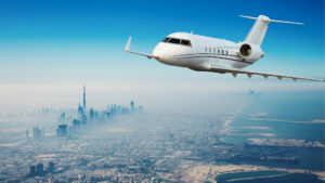 Ein weißer Privatjet fliegt über eine moderne Stadtlandschaft in Dubai mit Wolkenkratzern wie dem Burj Khalifa und einer Küste unter einem strahlend blauen Himmel – eine Szene, die Millionäre zu schätzen wissen.
