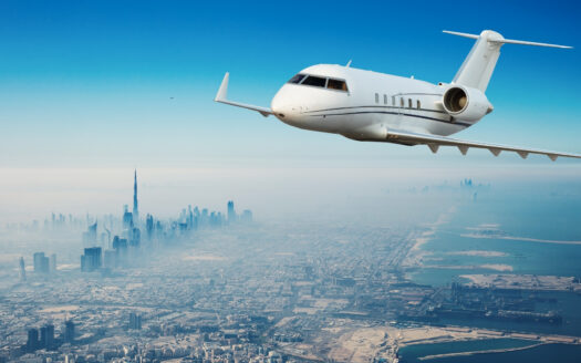 Un jet privé blanc survole un paysage urbain moderne à Dubaï, avec des gratte-ciel comme le Burj Khalifa et un littoral en contrebas, sous un ciel bleu clair - une scène appréciée des millionnaires.