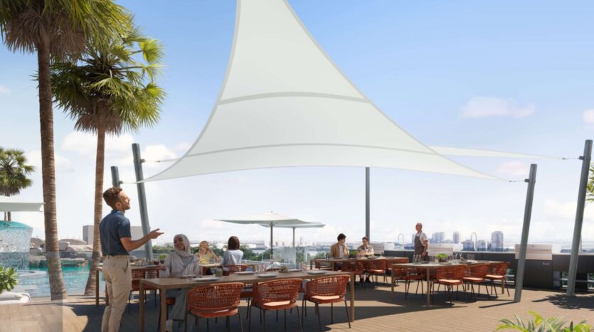 People are dining outdoors on the rooftop terrace of Alta View, by the pool, in the shade of a large white awning. Palm trees and city buildings rise in the background under a clear blue sky.