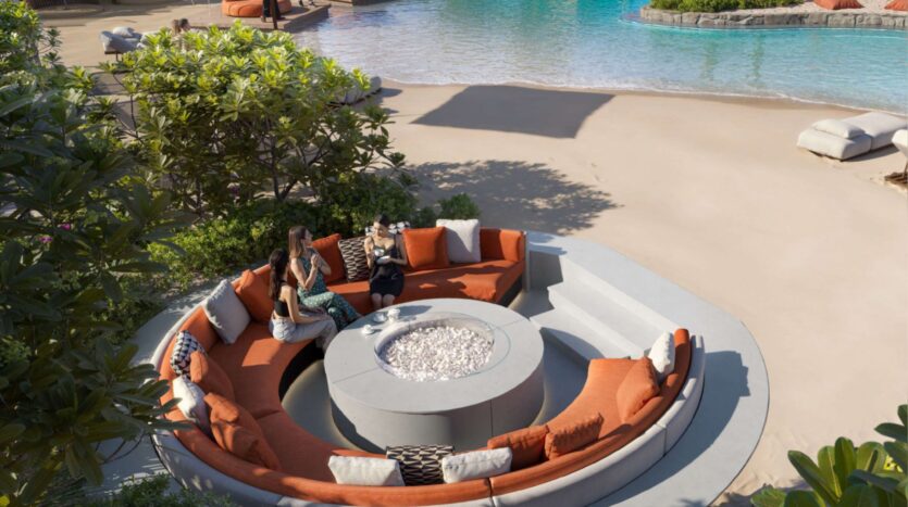 Two people sit and chat on an orange circular outdoor sofa at Alta View, around a fire pit next to a swimming pool. Tropical plants, sandy surfaces, and decorative cushions complete this welcoming space.