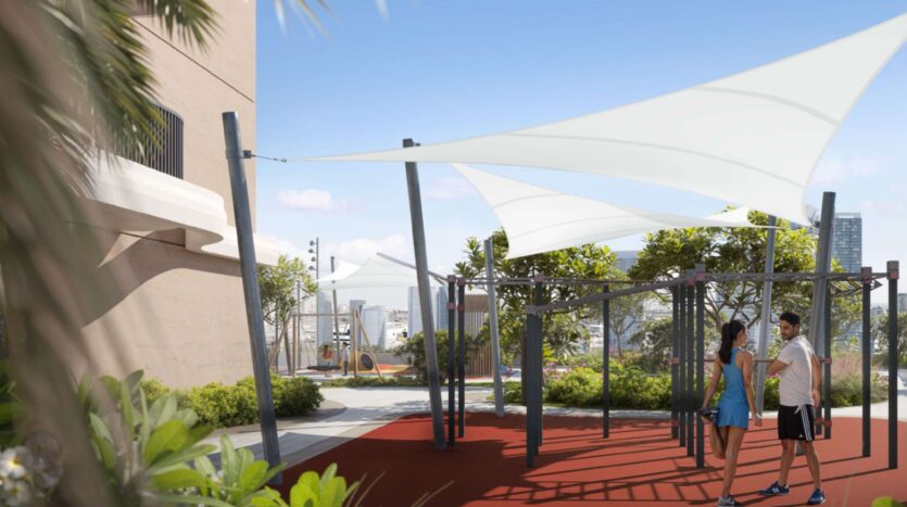 Two people stand and chat under shade sails in the outdoor exercise area at Alta View, surrounded by trees and plants on a sunny day, with the city buildings visible in the background.