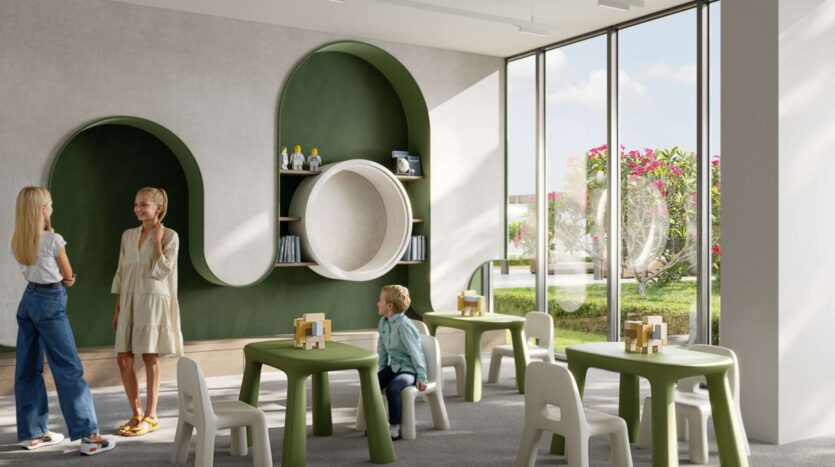 A modern, bright playroom for children at Alta View features green tables and white chairs. Two women stand talking while a child sits at a table. Shelves containing toys and books line a green wall, and large windows let in sunlight.