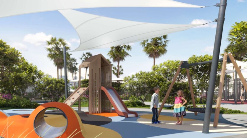 Two children stand and chat in the colorful Alta View playground, which features slides, swings, climbing equipment, and shade sails. Palm trees and greenery surround the area, all under a clear, sunny sky.
