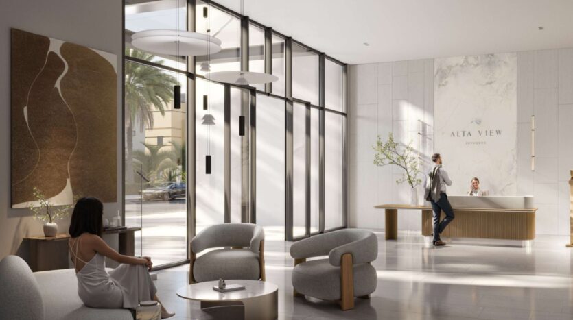 The modern and elegant lobby of the Alta View features large windows, minimalist furniture, artwork on the walls, and two people at the reception desk. A woman in a white dress is sitting on a chair while a receptionist assists a customer.