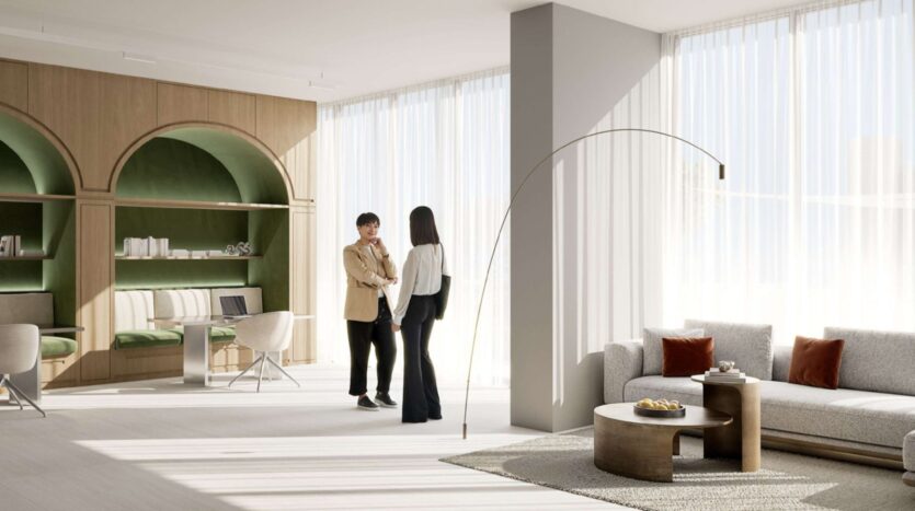 Two people are conversing in a modern, sunlit living room at Alta View, featuring large windows, a gray sofa, round coffee tables, and built-in shelves in a green alcove with a sitting area in the background.
