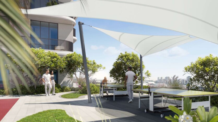 People play ping-pong under a white canopy on a shaded outdoor terrace at Alta View, surrounded by greenery and with the city skyline in the background. Two people walk nearby, adding to the bright and modern atmosphere of the scene.