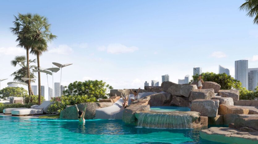 A person dives into a turquoise pool surrounded by rocks and greenery at Alta View, complete with a waterfall and slides. Modern urban buildings and palm trees are visible in the background under a clear blue sky.