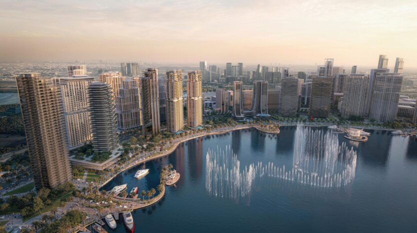 Aerial view of a modern city's waterfront showcasing the artistry of large glass buildings, boats moored in a marina, palm trees, and water fountains spraying a circular pattern onto a large body of water at sunset.