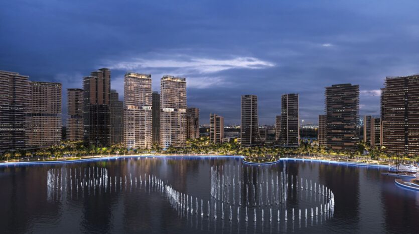 A nighttime cityscape shows modern skyscrapers surrounding a waterfront where illuminated fountains display artistry, creating patterns on the water, all under a cloudy blue sky.
