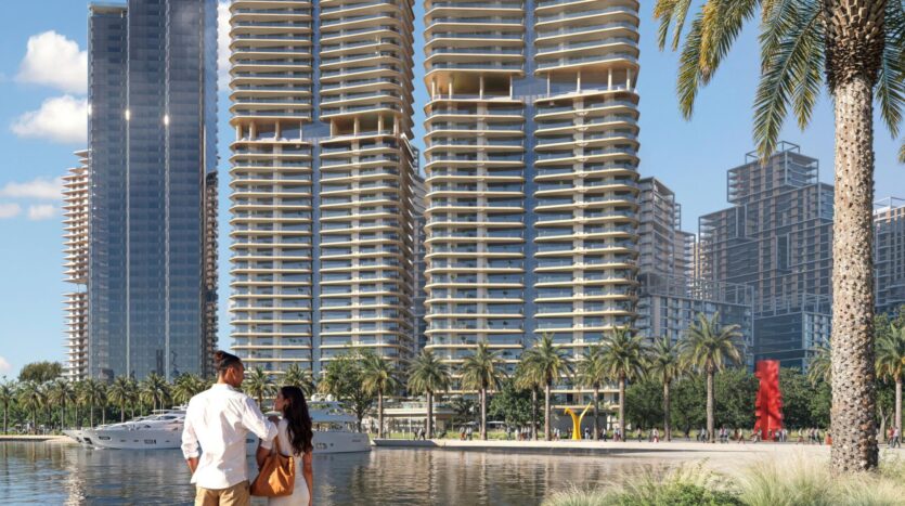 A couple stands at the water's edge, admiring the modern skyscrapers with rounded balconies, palm trees, nearby moored yachts, and vibrant public art sculptures that showcase the city's artistry in a sunny urban setting.