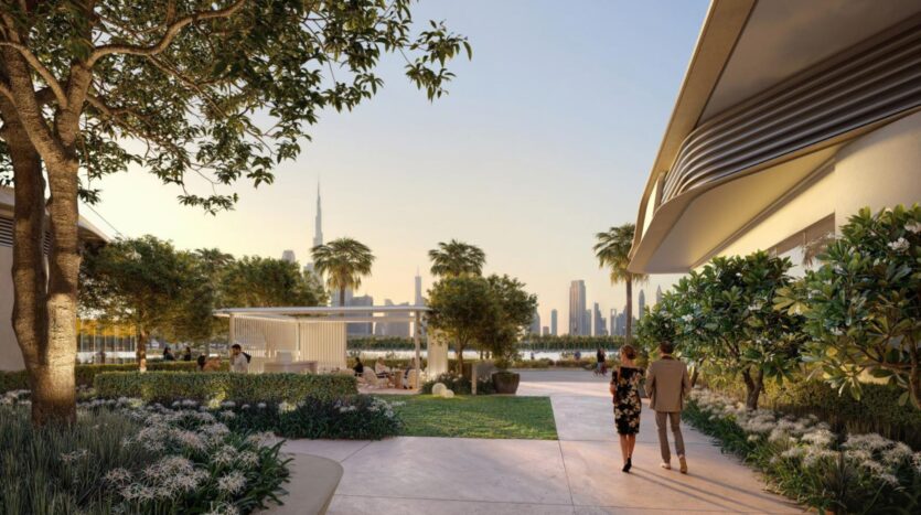 A couple walks along a path in a modern park, surrounded by lush vegetation and palm trees. The city's skyscrapers, including the Burj Khalifa, rise up on the horizon at sunset.