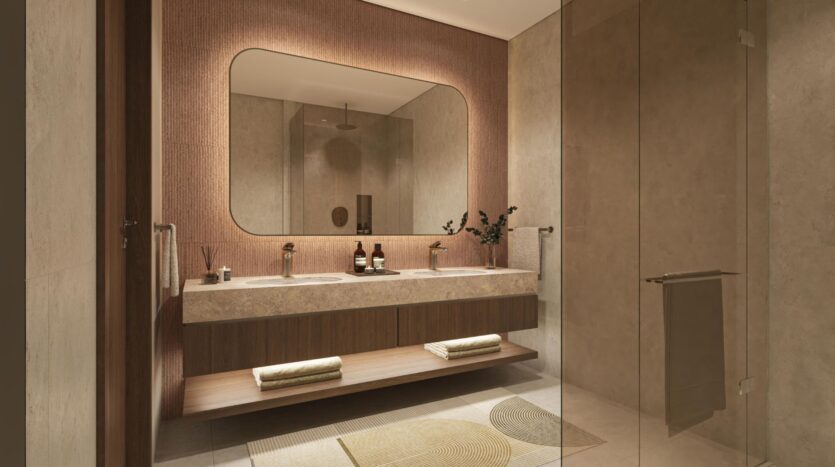 A modern bathroom that showcases art with a large mirror, double sink, brown wood and stone finishes, towels folded on a shelf, a glass shower stall, and a decorative rug on light-colored tile flooring.