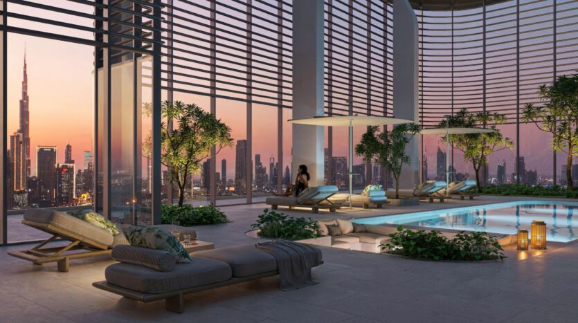 The luxurious indoor pool, complete with lounge chairs, modern décor, and touches of greenery, showcases art, while large windows frame a breathtaking city skyline at sunset; a woman relaxes on a chair near the sparkling pool.