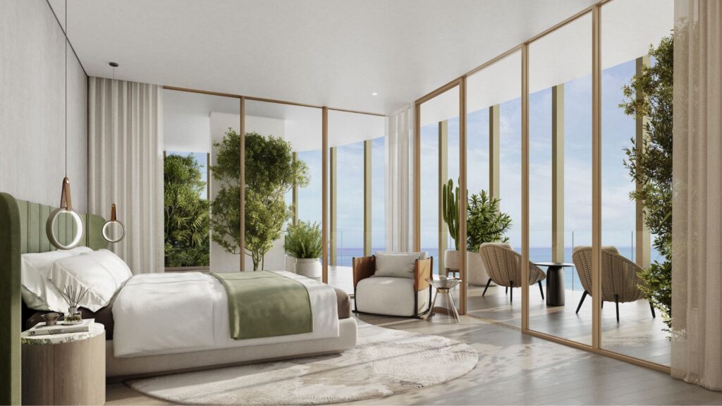 A modern Kanyon room with floor-to-ceiling windows, a large bed with green accents, a comfortable sitting area, indoor plants, and a balcony with wicker chairs overlooking the ocean. The space is bathed in natural light.