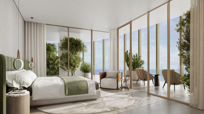 A modern Kanyon room with floor-to-ceiling windows, a large bed with green accents, a comfortable sitting area, indoor plants, and a balcony with wicker chairs overlooking the ocean. The space is bathed in natural light.