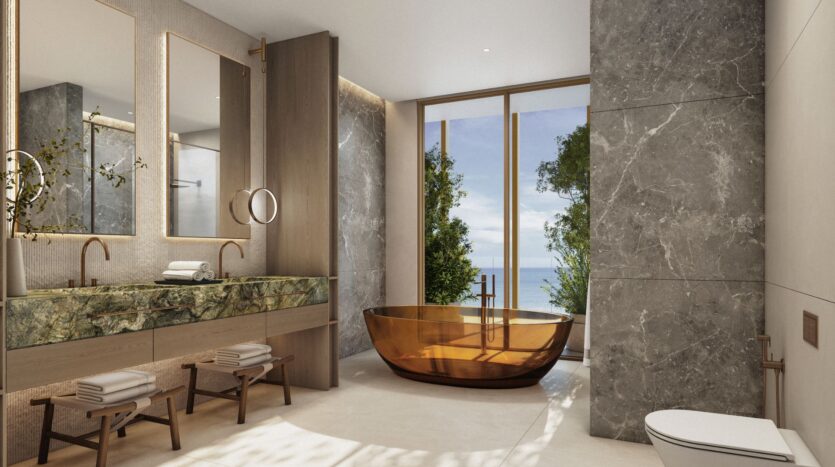 Luxurious modern bathroom with a large marble wall, two sinks, mirrors, and a transparent amber Kanyon bathtub, floor-to-ceiling windows, white tile flooring, and breathtaking views of the trees and ocean outside.