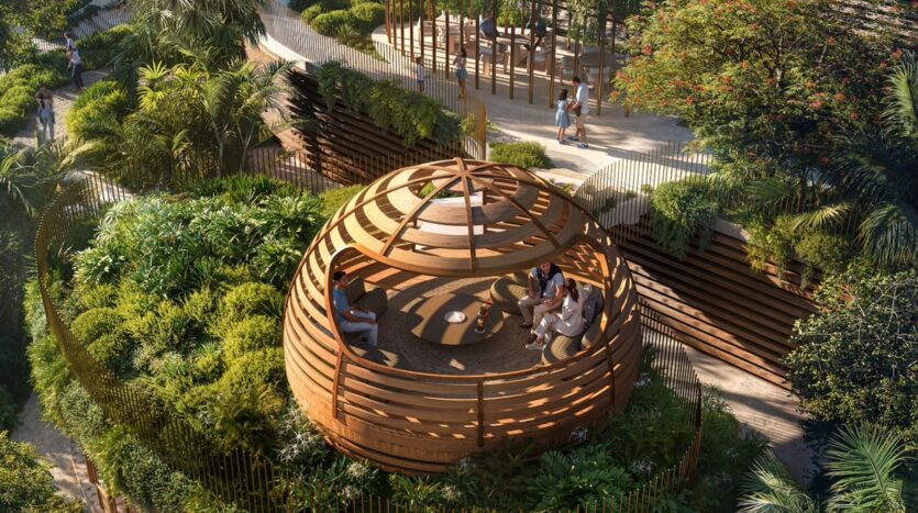 A group of people are sitting and chatting inside a round, open wooden gazebo in Kanyon, surrounded by lush greenery and trees in a landscaped garden. Sunlight filters through the slatted roof, creating a natural and peaceful setting.