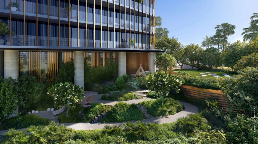 A modern building with large bay windows overlooks a lush landscaped garden at Kanyon, featuring winding paths, various green plants, trees, and a small open space to sit under a clear blue sky.