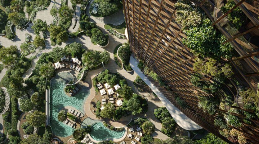 Aerial view of a lush garden in Kanyon, featuring winding paths, swimming pools, lounge chairs, and abundant greenery next to a large building covered in plants.