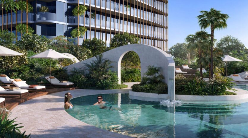 Kanyon's modern outdoor pool features a curved white deck, lush greenery, palm trees, lounge chairs, and umbrellas. Two people swim and relax in the clear blue water.