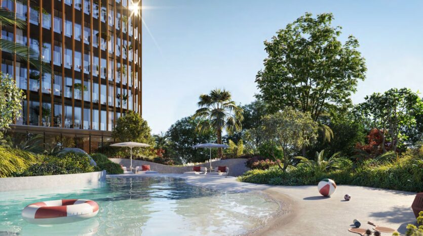 A modern apartment building by Kanyon overlooks a resort-style swimming pool with lifebuoys, lush landscaping, sun loungers, and parasols under a clear blue sky. Sunlight reflects off the windows and water, enhancing the relaxing atmosphere.