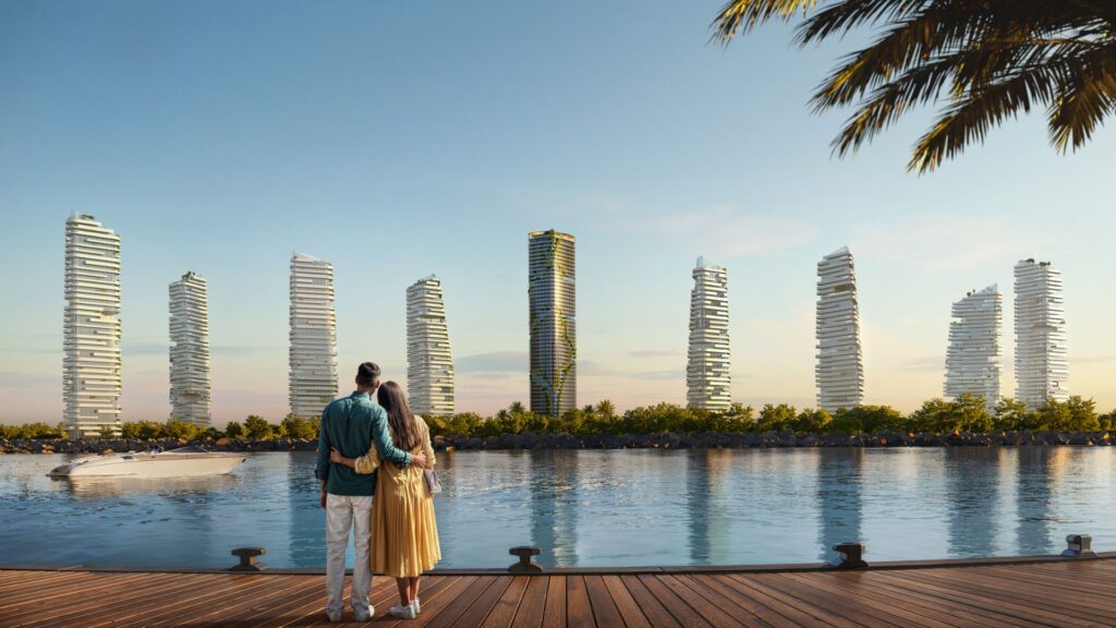 A couple stands on a wooden pier at the water's edge, gazing at a skyline of uniquely shaped modern skyscrapers, such as Kanyon, with palm trees and a boat in the background, under clear skies.