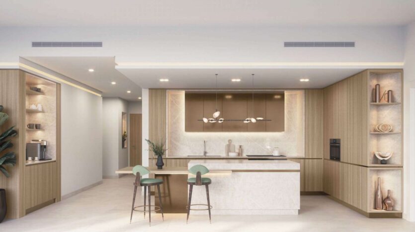 Cuisine moderne avec des armoires en bois clair, un îlot en marbre avec deux tabourets de bar verts, des étagères encastrées avec de la décoration, des lampes suspendues et des murs aux tons neutres. L'espace inspiré de Soto Grande est lumineux et minimaliste.