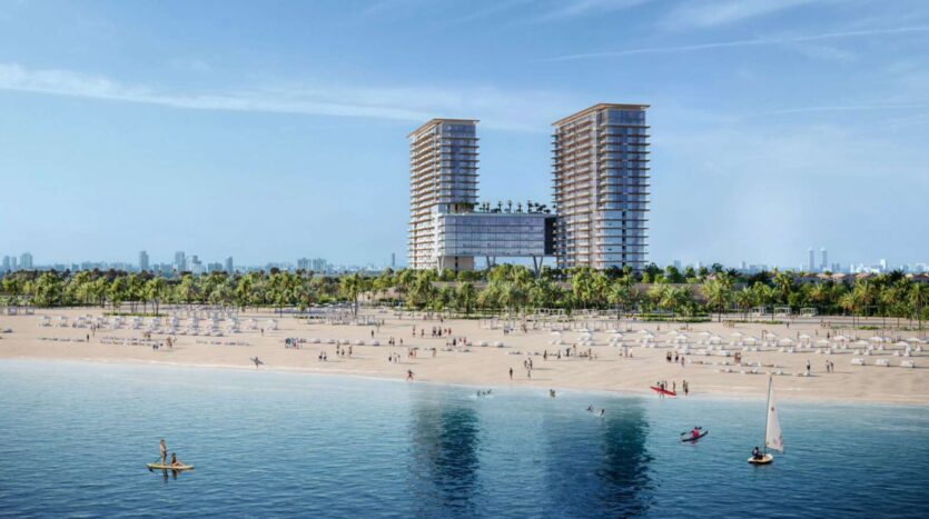 Une grande plage de sable à Soto Grande avec des gens qui se promènent et se détendent près de l'eau, bordée de palmiers. À l'arrière-plan, deux grands bâtiments modernes avec une structure de liaison se détachent sur le ciel bleu, tandis que de petits bateaux et des kayaks sillonnent l'eau.