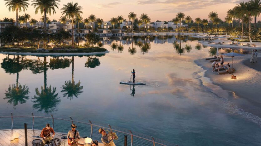 A serene waterside scene at sunset in The Heights, featuring a person paddleboarding on calm waters, people relaxing on a sandy beach, and musicians playing on a wooden deck surrounded by palm trees and modern villas.
