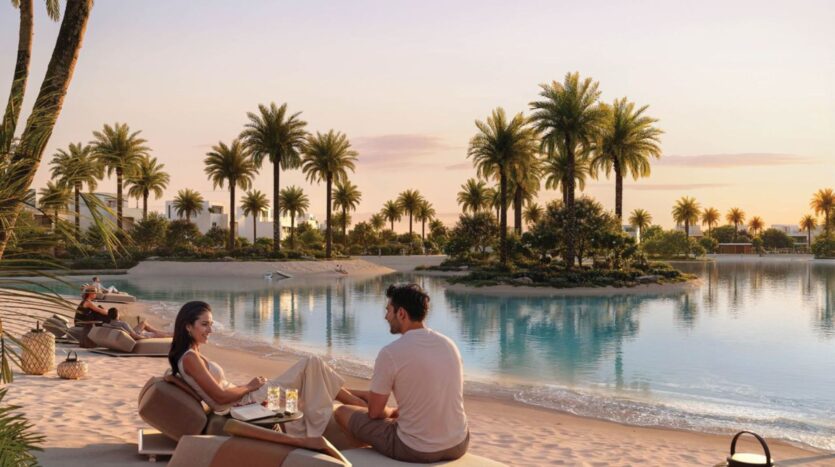 A man and a woman relax on lounge chairs by a tranquil lagoon at sunset in The Heights, surrounded by palm trees and a sandy beach. Drinks and a bag are placed nearby, while others lounge in this peaceful, tropical atmosphere.