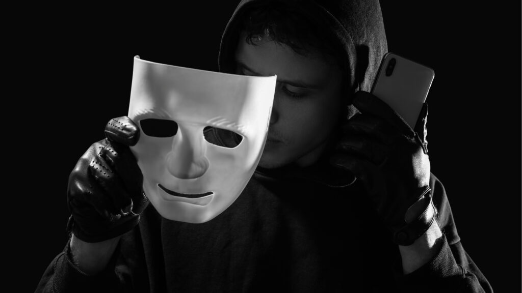 Una persona che indossa una felpa con cappuccio e guanti neri tiene una maschera bianca in una mano e uno smartphone nell'altra, nascondendo parzialmente il proprio volto, a simboleggiare gli investitori che cercano di evitare le truffe immobiliari a Dubai su uno sfondo scuro.