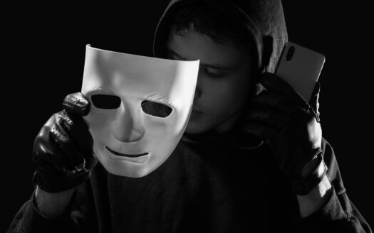 Une personne portant un sweat à capuche et des gants noirs tient un masque blanc dans une main et un smartphone dans l'autre, masquant partiellement son visage, symbolisant les investisseurs cherchant à éviter les arnaques immobilières Dubaï sur un fond sombre.