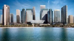 Eine moderne Stadtlandschaft von Abu Dhabi mit hohen Glaswolkenkratzern und einzigartigen architektonischen Entwürfen, darunter die Erweiterung des ADGM, spiegelt sich in einem ruhigen Gewässer unter blauem Himmel wider.