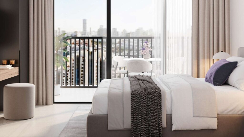 A modern Alta View room with a neatly made bed, gray and white bedding, a purple pillow, and a black throw blanket. Large glass doors open onto a balcony with a table, chairs, and a stunning view of the city in the background.