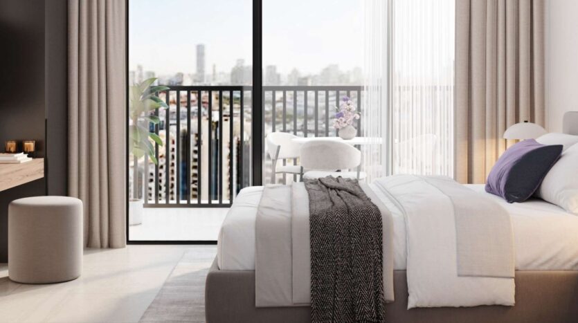 A modern Alta View room with a neatly made bed, gray and white bedding, a purple pillow, and a black throw blanket. Large glass doors open onto a balcony with a table, chairs, and a stunning view of the city in the background.