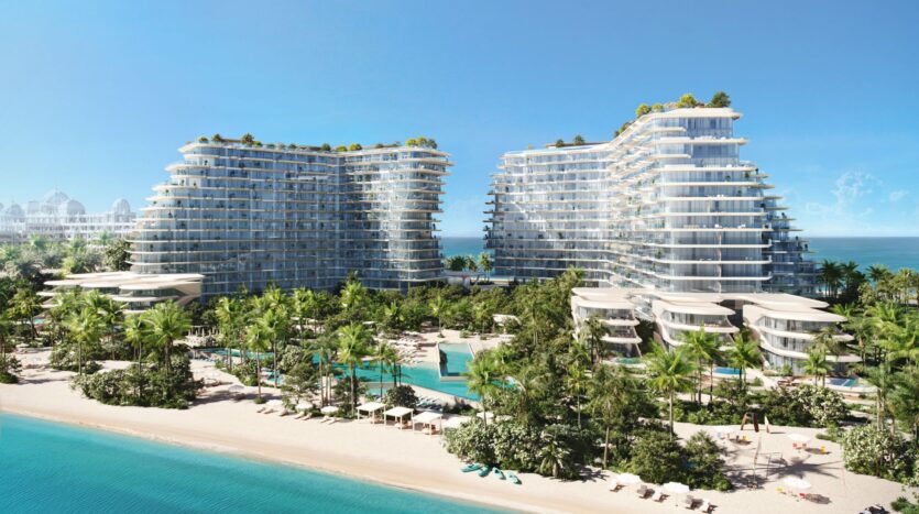 The two modern, glass-fronted towers of Bella by Passo rise above palm trees and gardens, overlooking a beautiful white sand beach and turquoise water. Swimming pools, parasols, and sun loungers are set up near the shore under a clear blue sky.
