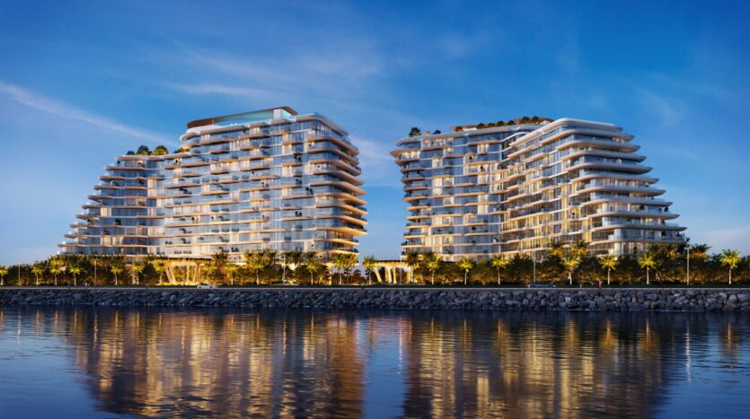 At Bella by Passo, two modern multi-story residential buildings with curved balconies stand side by side, illuminated at dusk and reflected in the calm waters of the seafront, surrounded by palm trees.
