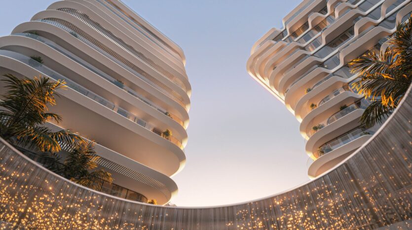 Low-angle view of two modern, white, curved buildings with balconies at Bella by Passo, palm trees, and a circular structure below decorated with twinkling lights, all set against a clear sky at sunset.