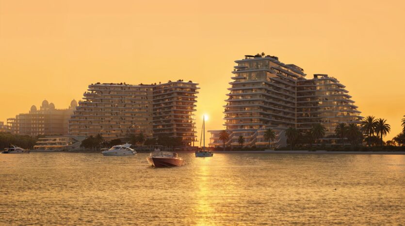 Sunset over the modern terraced buildings of Bella by Passo, at the water's edge, with boats anchored in calm waters. The sky is orange and palm trees line the shore, reflecting a peaceful and luxurious atmosphere.
