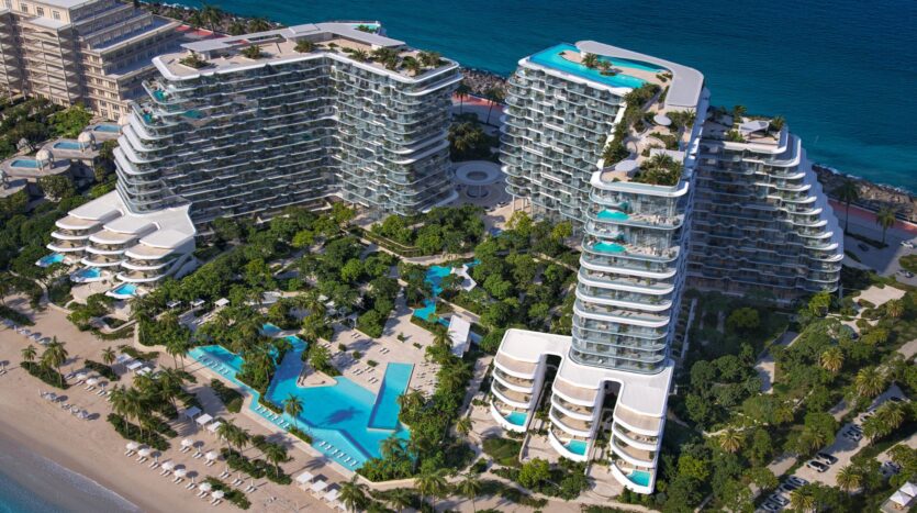 Aerial view of Bella by Passo, a large modern seaside complex with curved glass-walled buildings, numerous swimming pools, lush palm trees, and relaxation areas, all overlooking the turquoise ocean.