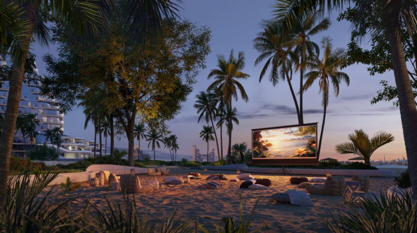 An outdoor cinema set up on a sandy beach at dusk is enhanced with touches from Bella by Passo—pillows and blankets to sit on, surrounded by palm trees. A large screen displays a sunset scene with modern buildings in the background.