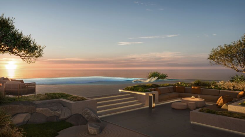 The luxurious outdoor lounge at Bella by Passo features a sunken seating area and fireplace, overlooking the infinity pool and calm ocean at sunset, surrounded by lush greenery and modern décor.