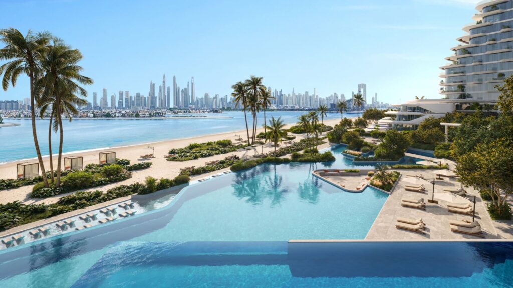 Infinity pool overlooking a sandy beach with palm trees, Bella by Passo luxury cabanas, and a modern city in the background under a clear blue sky. Sun loungers are arranged next to the pool.