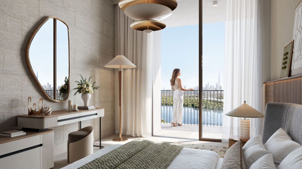 Dormitorio moderno con tonos neutros, una cama grande y un escritorio con espejo. Una mujer se encuentra en el balcón exterior, que da a Creek Bay y al horizonte de la ciudad a través de puertas acristaladas que van del suelo al techo. La luz natural inunda este elegante espacio.