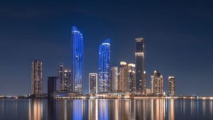 Die Skyline einer modernen Stadt bei Nacht mit großen, beleuchteten Wolkenkratzern und blauen und weißen Lichtern, die sich im ruhigen Wasser der Creek Bay im Vordergrund spiegeln. Der Himmel ist dunkel und klar.