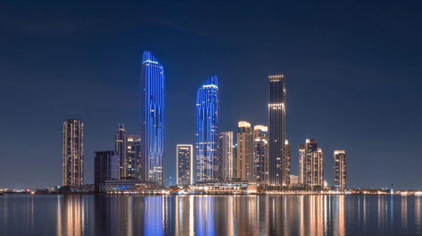 El horizonte nocturno de una ciudad moderna, con grandes rascacielos iluminados y luces azules y blancas reflejadas en las tranquilas aguas de la bahía de Creek en primer plano. El cielo está oscuro y despejado.